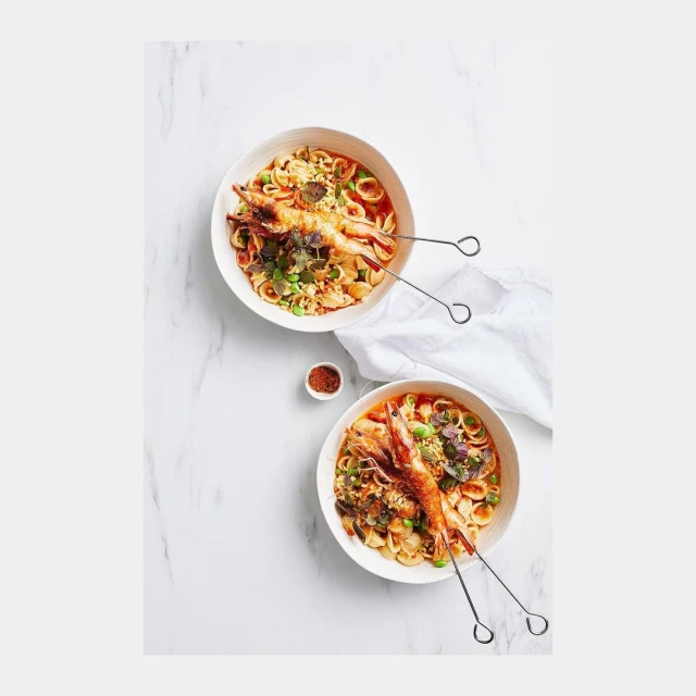 Overhead shot of two bowls of pasta with shrimp and herbs on a white marble surface with a small dish of seasoning.