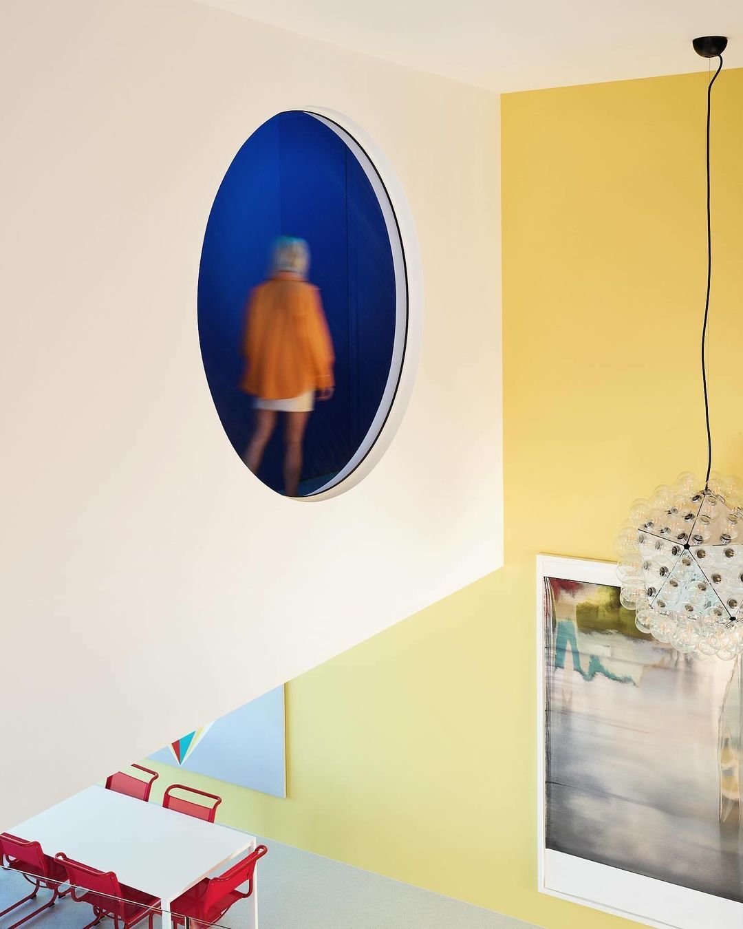 Interior shot of an abstract painting, modern lamp, yellow wall, red chairs, and a person behind a round window in the wall.