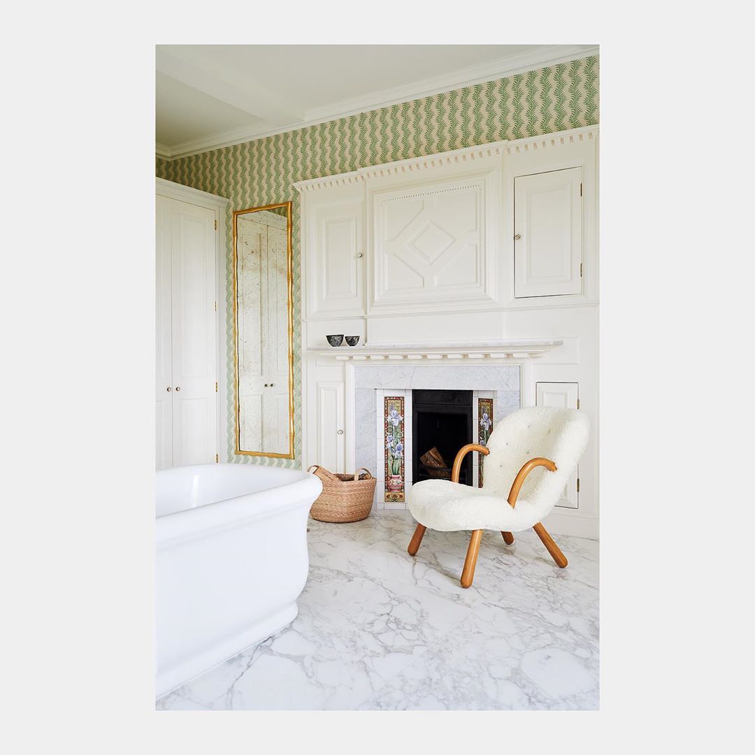 White freestanding bathtub and cream armchair in a luxurious bathroom with white marble floors and green patterned wallpaper