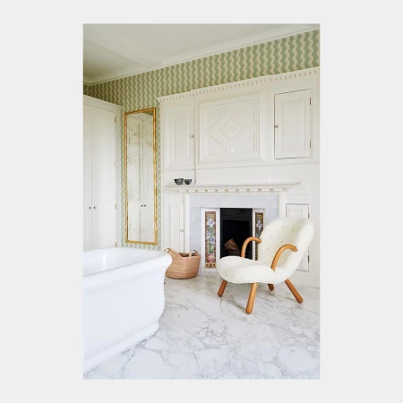 White freestanding bathtub and cream armchair in a luxurious bathroom with white marble floors and green patterned wallpaper