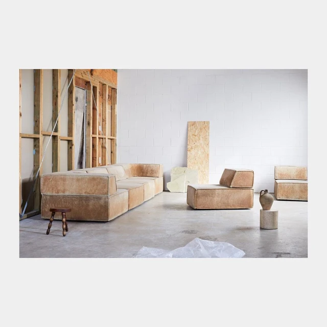 Living room interior showcasing a modular sofa, chair, and stool, with neutral colors and wood accents against a white brick wall.