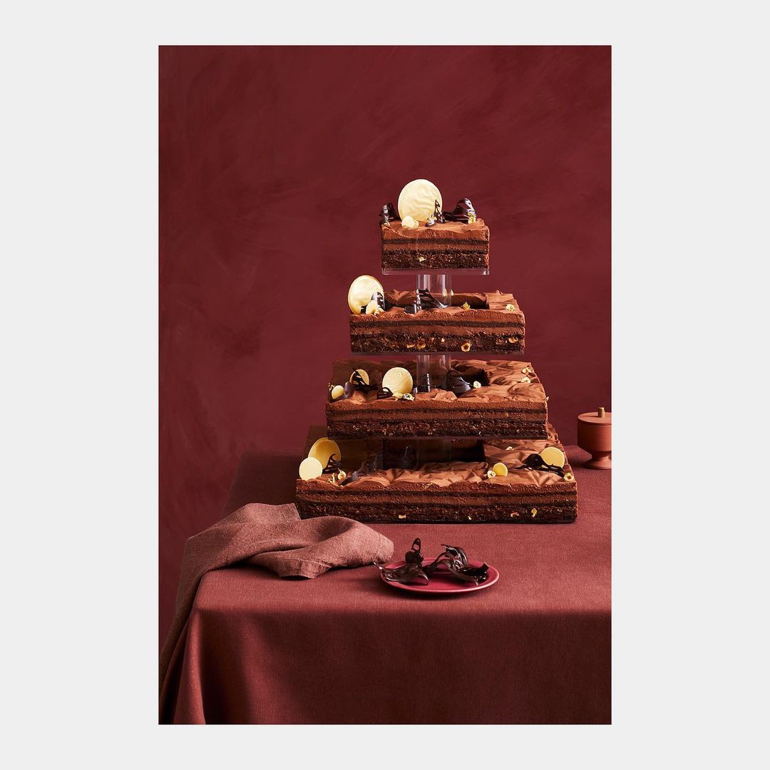 Three-tiered chocolate dessert tower with frosting, chocolate shavings and pieces, presented on a burgundy linen tablecloth.