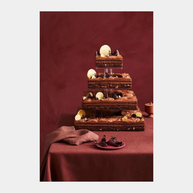 Three-tiered chocolate dessert tower with frosting, chocolate shavings and pieces, presented on a burgundy linen tablecloth.