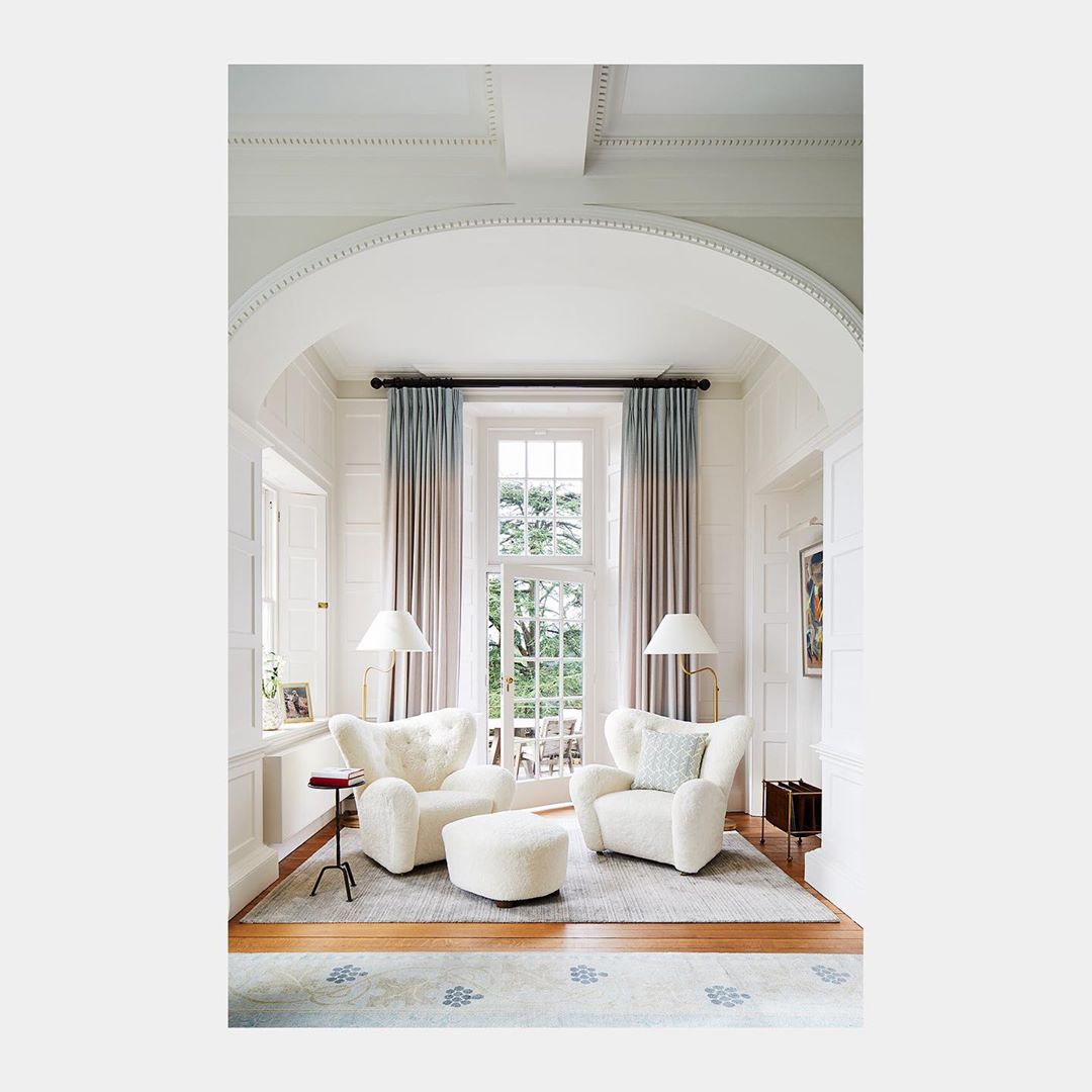 Living room interior features two white armchairs and an ottoman, complemented by floor lamps and curtains by the window.