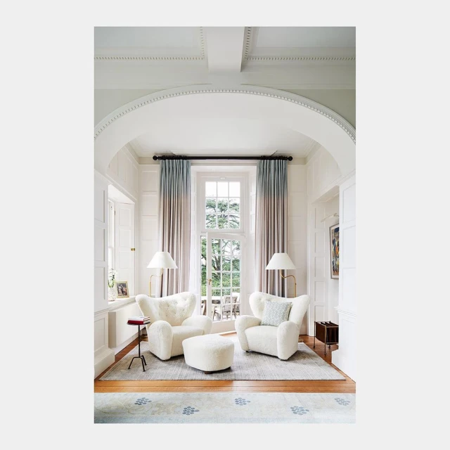 Living room interior features two white armchairs and an ottoman, complemented by floor lamps and curtains by the window.