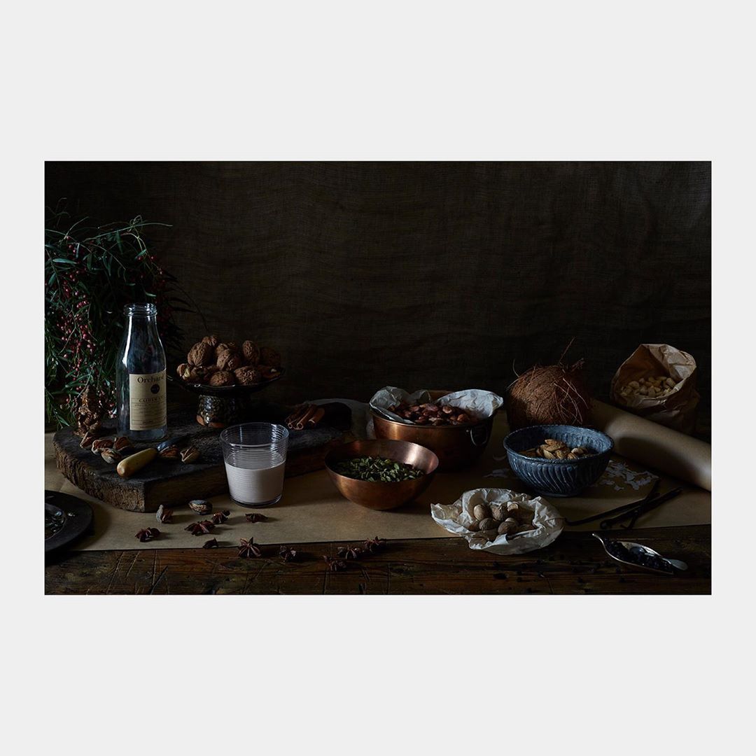 Still life of nuts, seeds, spices, and a beverage in glass containers, arranged on a wooden table against a dark background.