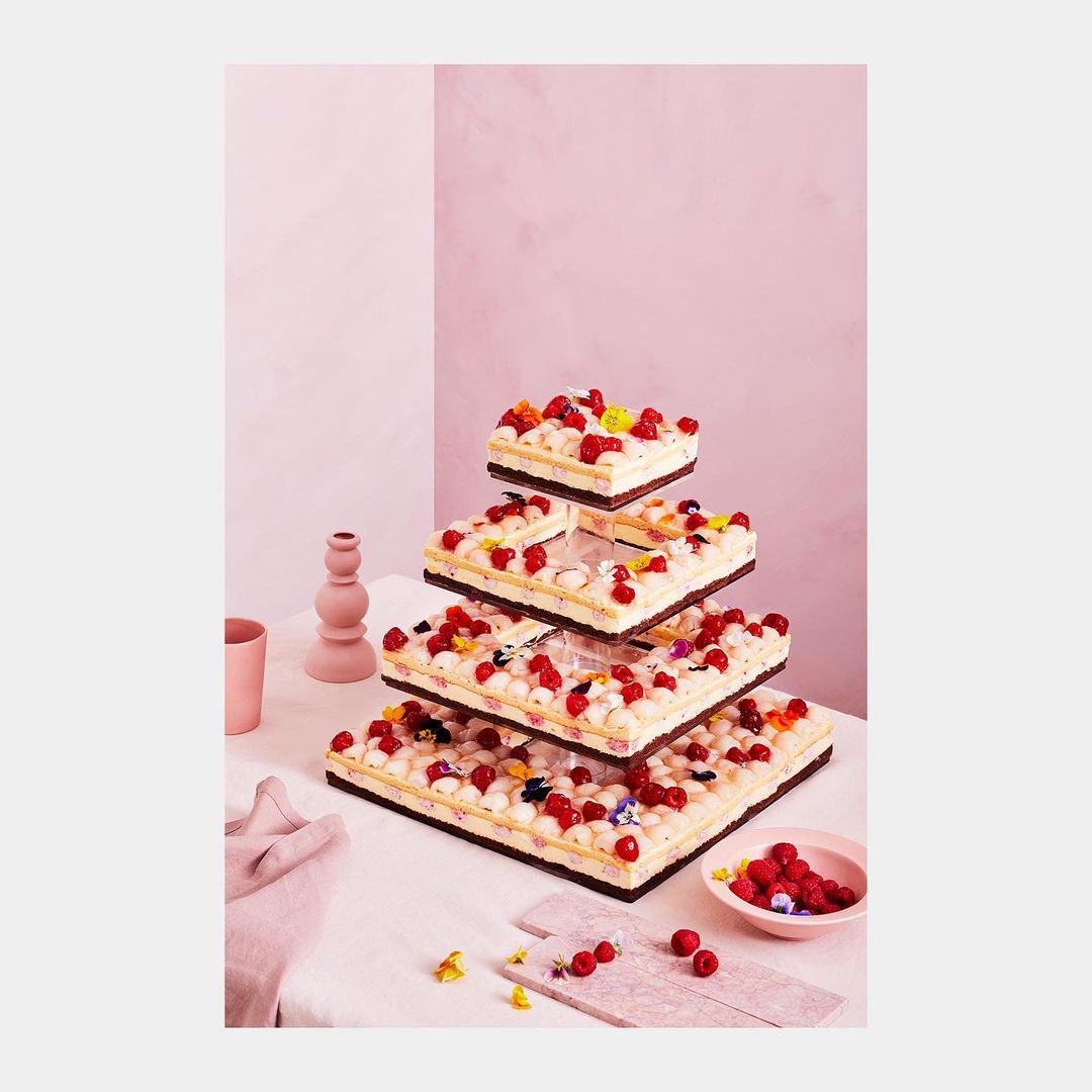 Square cake tower decorated with berries and flowers on a white table against a pink background, cake stand alternative, dessert.