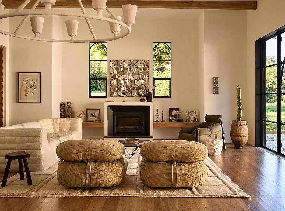 Living room interior with a white sofa, woven poufs, a fireplace, and geometric wall art, illuminated by natural light and a modern chandelier.