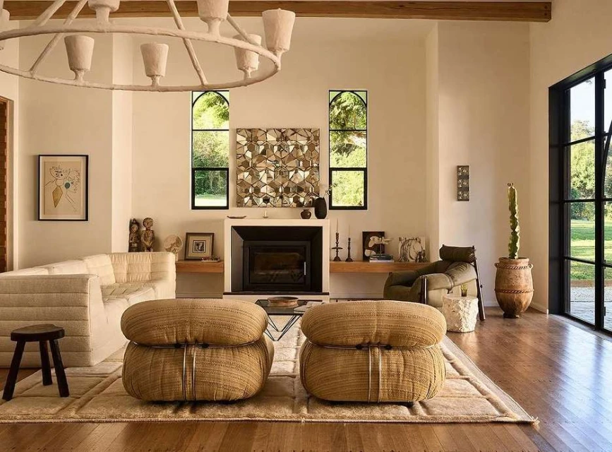 Living room interior with a white sofa, woven poufs, a fireplace, and geometric wall art, illuminated by natural light and a modern chandelier.
