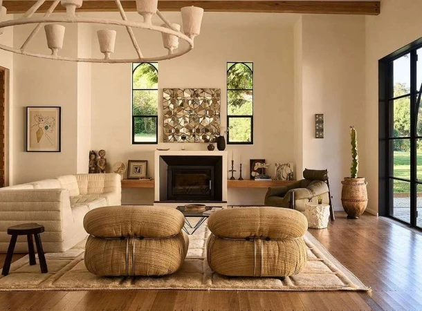 Living room interior with a white sofa, woven poufs, a fireplace, and geometric wall art, illuminated by natural light and a modern chandelier.