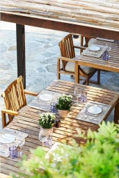Wooden outdoor dining tables set with plates, glassware, and potted plants under a thatched pergola in London by Alicia Taylor