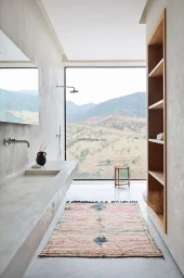 Minimalist modern bathroom with concrete vanity, built-in wooden shelving, patterned rug, and large window overlooking mountain landscape.