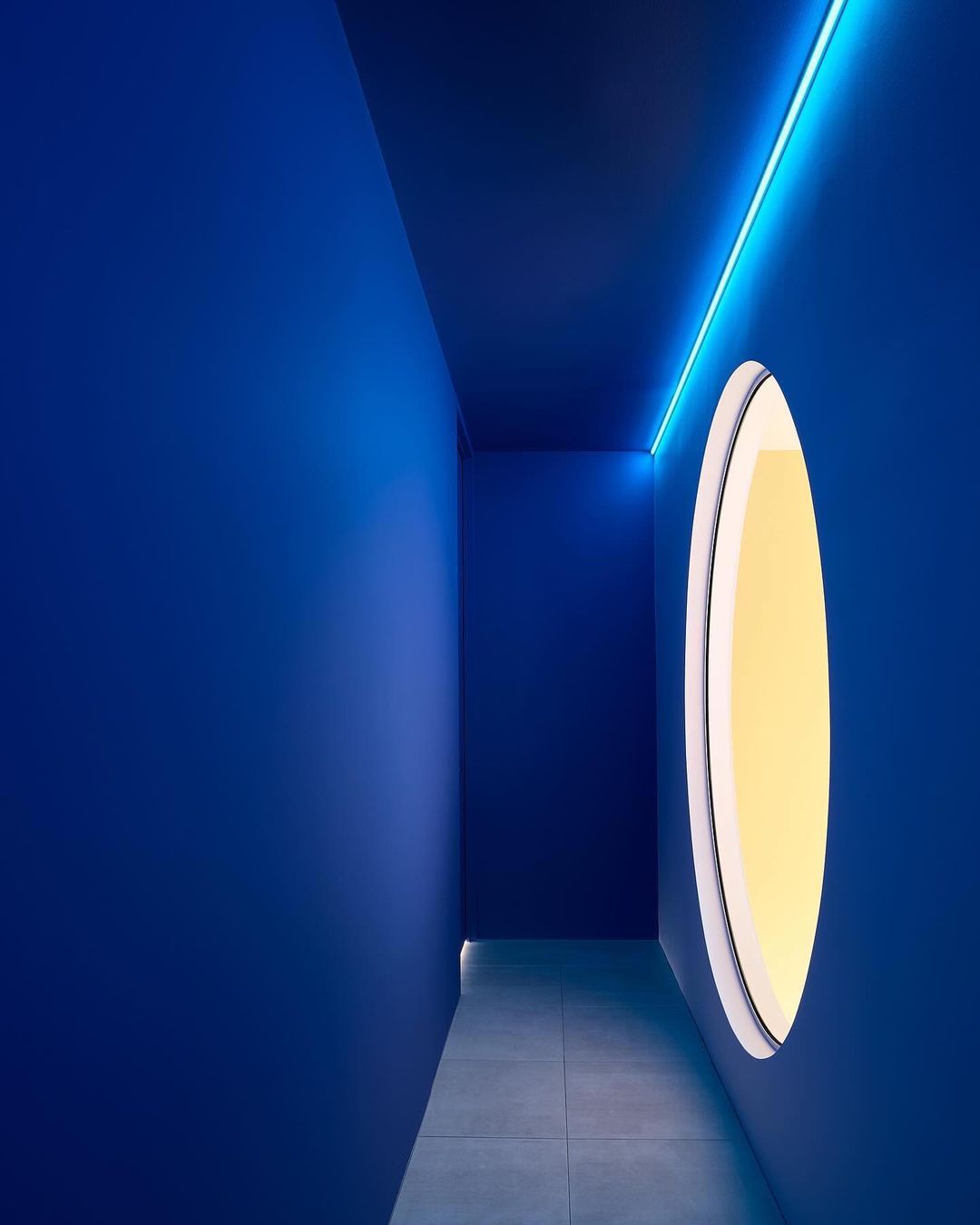 Narrow blue corridor with smooth walls and light grey tiled floor featuring a bright circular window on the right wall