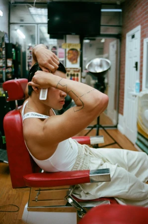 Young adult male in a barbershop chair, styling his hair with a comb. Red and white chair, visible Tommy Hilfiger branded straps.
