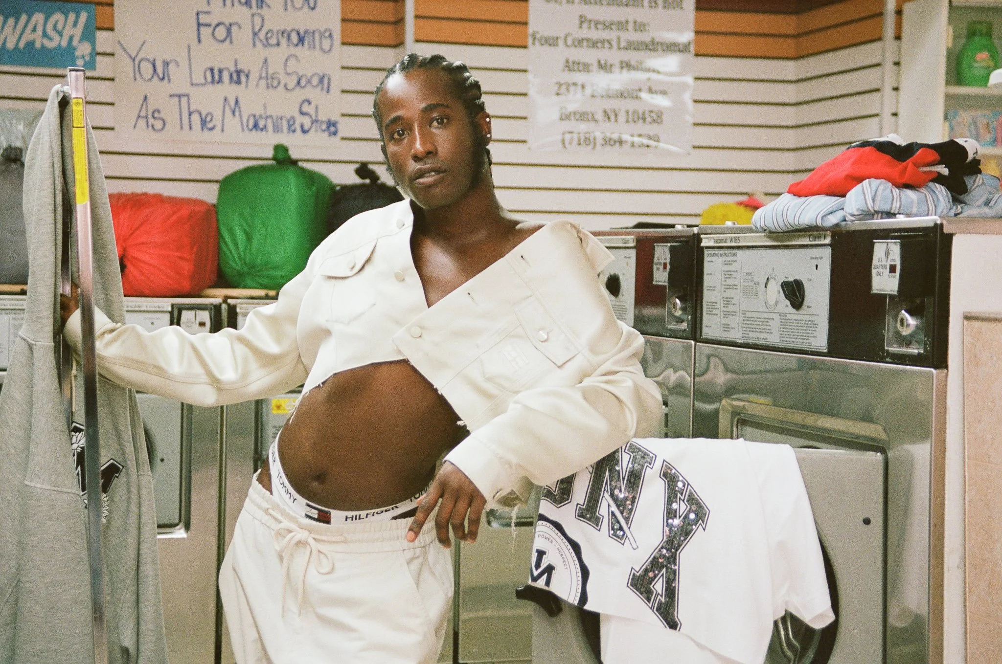 Adult male poses in a laundromat wearing white outfit; t-shirts and washers are visible. Neutral color scheme with fluorescent lighting.