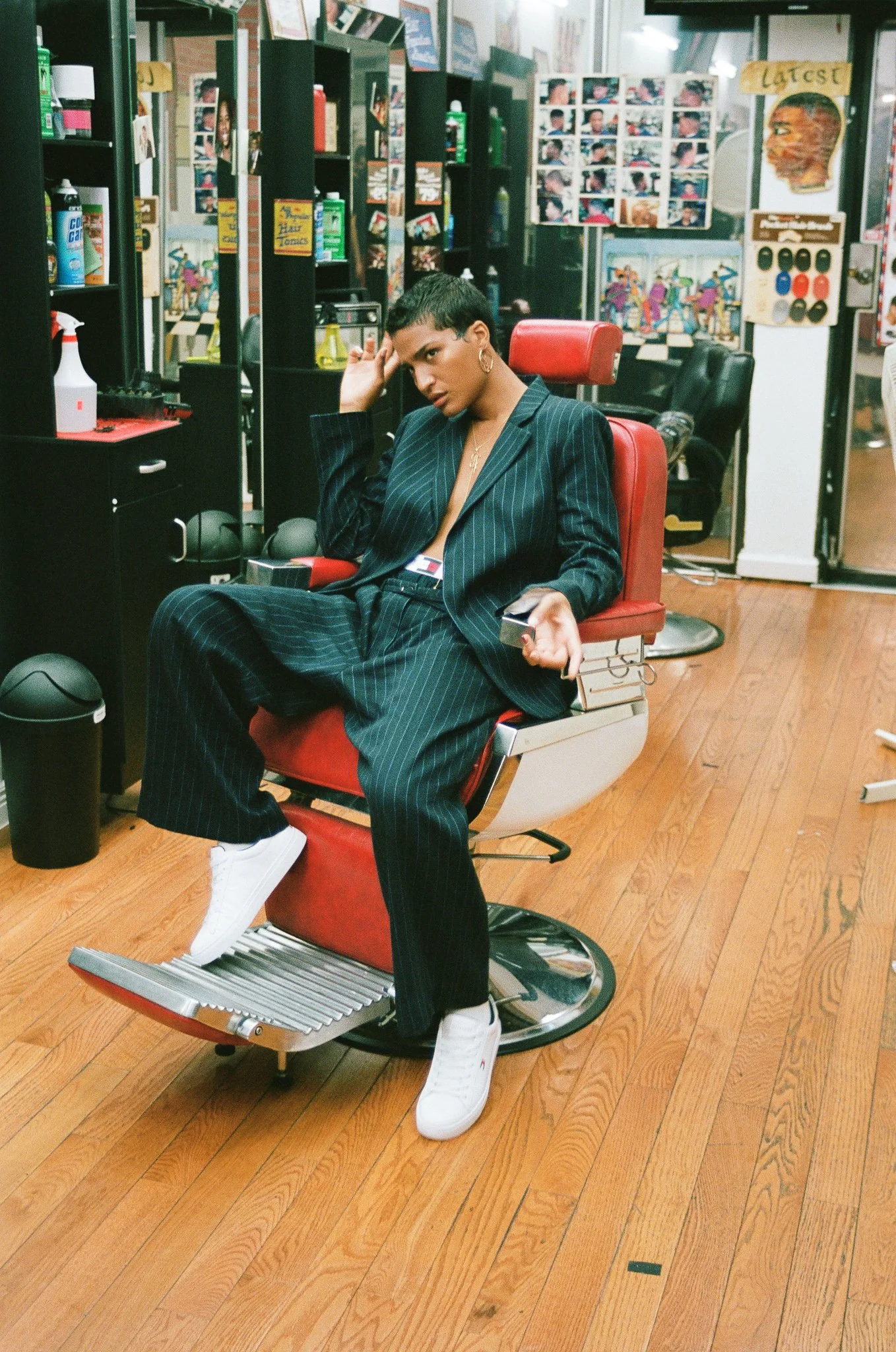 Man in pinstripe suit and white sneakers reclines in red barber chair in vintage-style barbershop with white-tiled walls, dark cabinets.