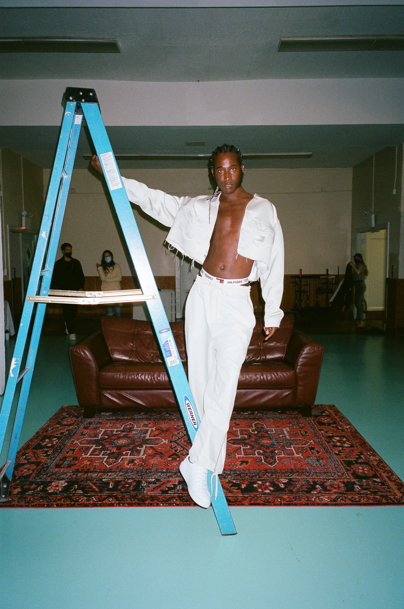 An adult male poses on a light-blue aluminum ladder in a photo studio. He is wearing a white jacket, trousers, and sneakers.