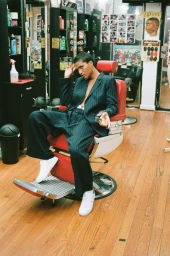 Person with short hair wearing a striped suit seated in a red barber chair inside a barbershop
