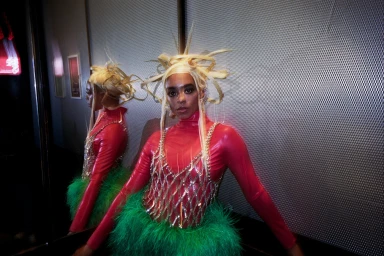 Woman with sculptural blonde hair wearing a red latex top, crystal harness overlay, and green feather skirt against a metal wall.