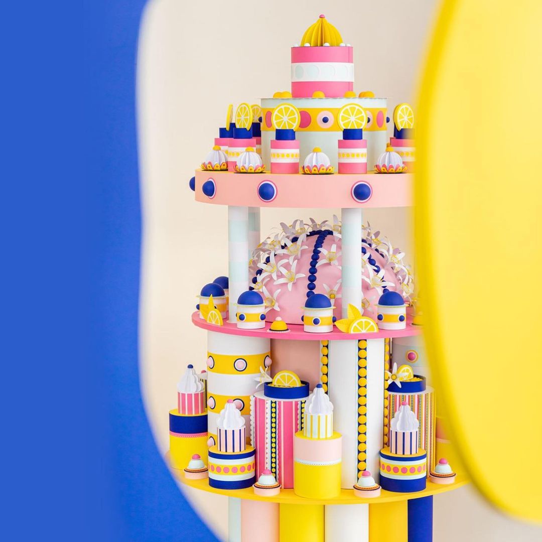 Three-tier whimsical cake with lemon accents, set against a backdrop of blue, white, and yellow curves in an indoor setting.