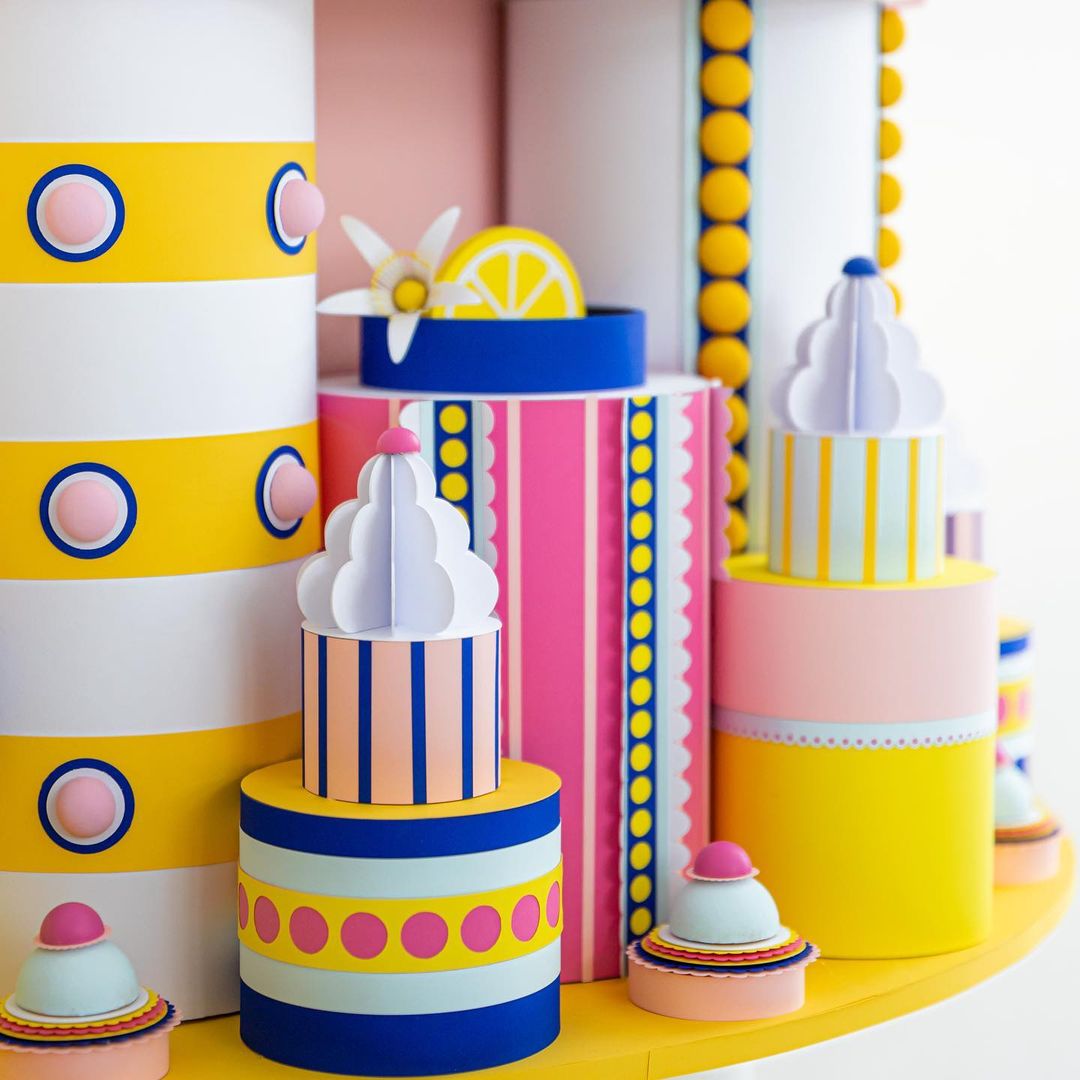 Colorful paper art featuring cylinder and cupcake shapes with stripes, polka dots, and pastel hues, arranged on a yellow surface.
