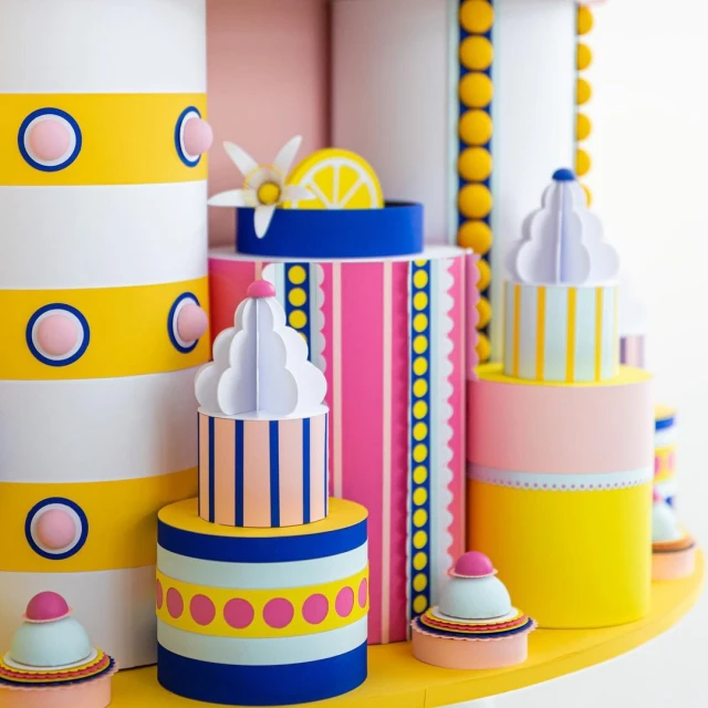 Colorful paper art featuring cylinder and cupcake shapes with stripes, polka dots, and pastel hues, arranged on a yellow surface.