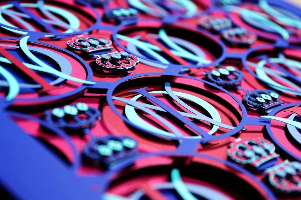 Intricate paper art featuring layers of blue, white, and red, creating abstract circular designs and crown shapes in a detailed pattern.