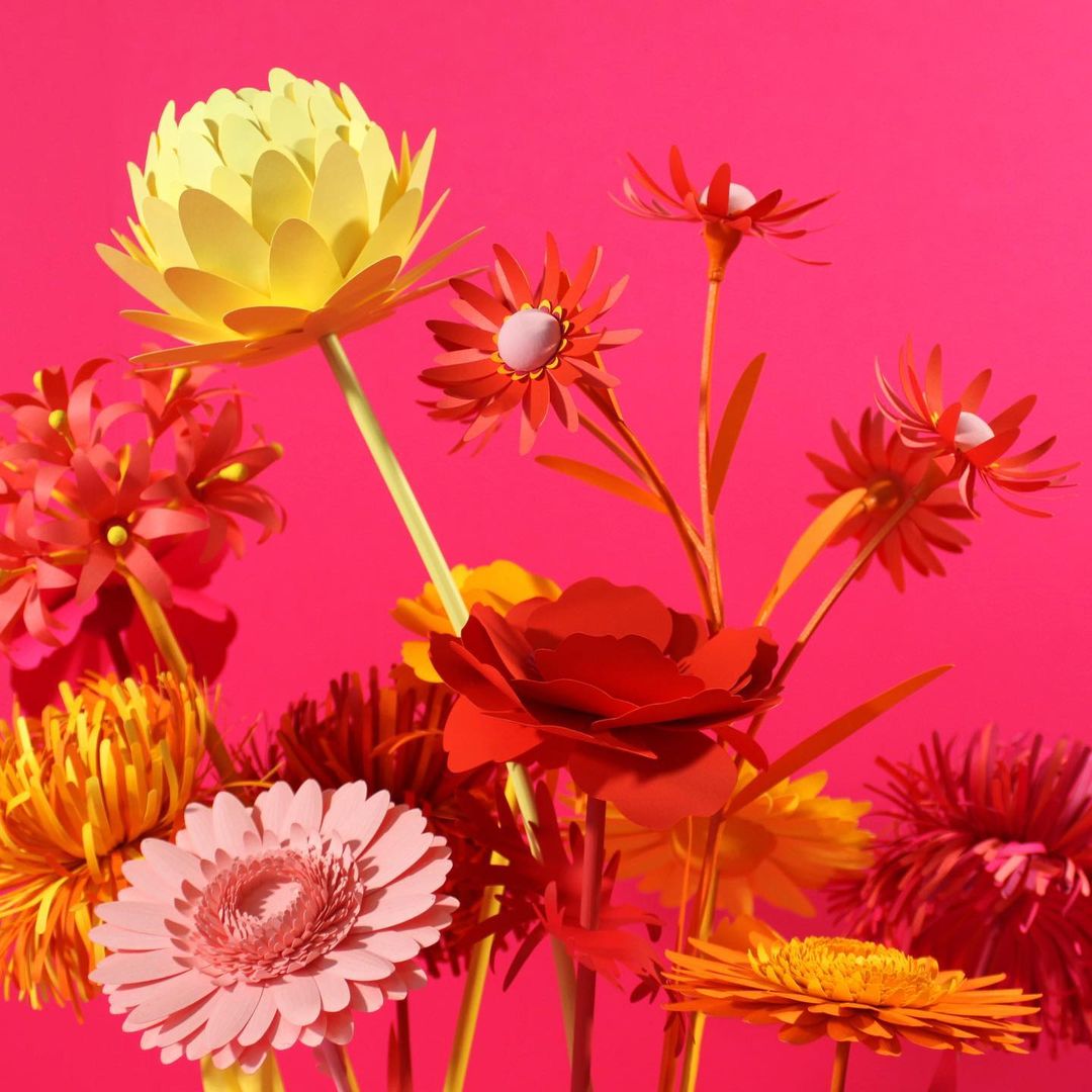 Vibrant paper flowers in shades of yellow, pink, and red against a bright pink background, showcasing a colorful floral display.