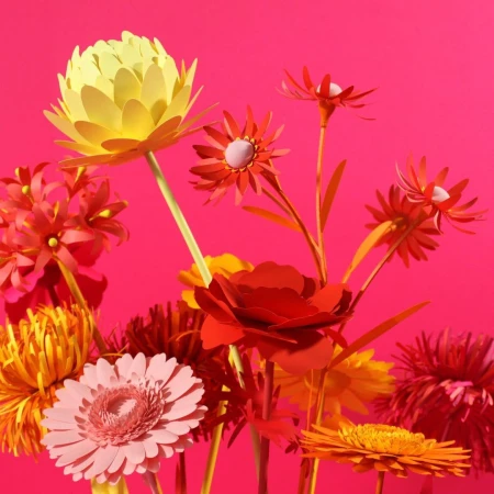 Vibrant paper flowers in shades of yellow, pink, and red against a bright pink background, showcasing a colorful floral display.