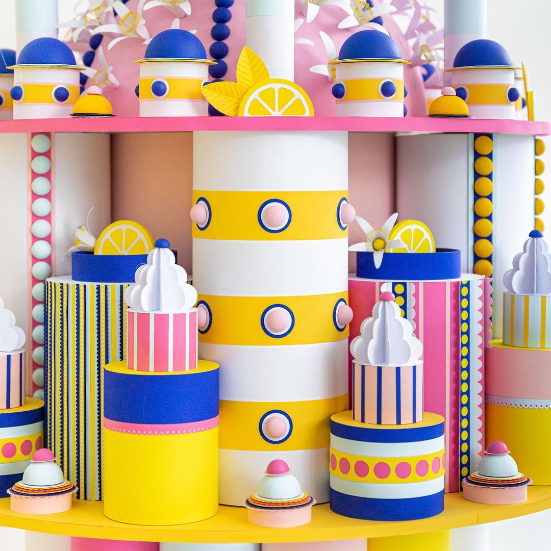 Colorful, whimsical still life of cake and lemon slice paper art with round geometric shapes and vibrant yellow, pink, blue, and white hues.