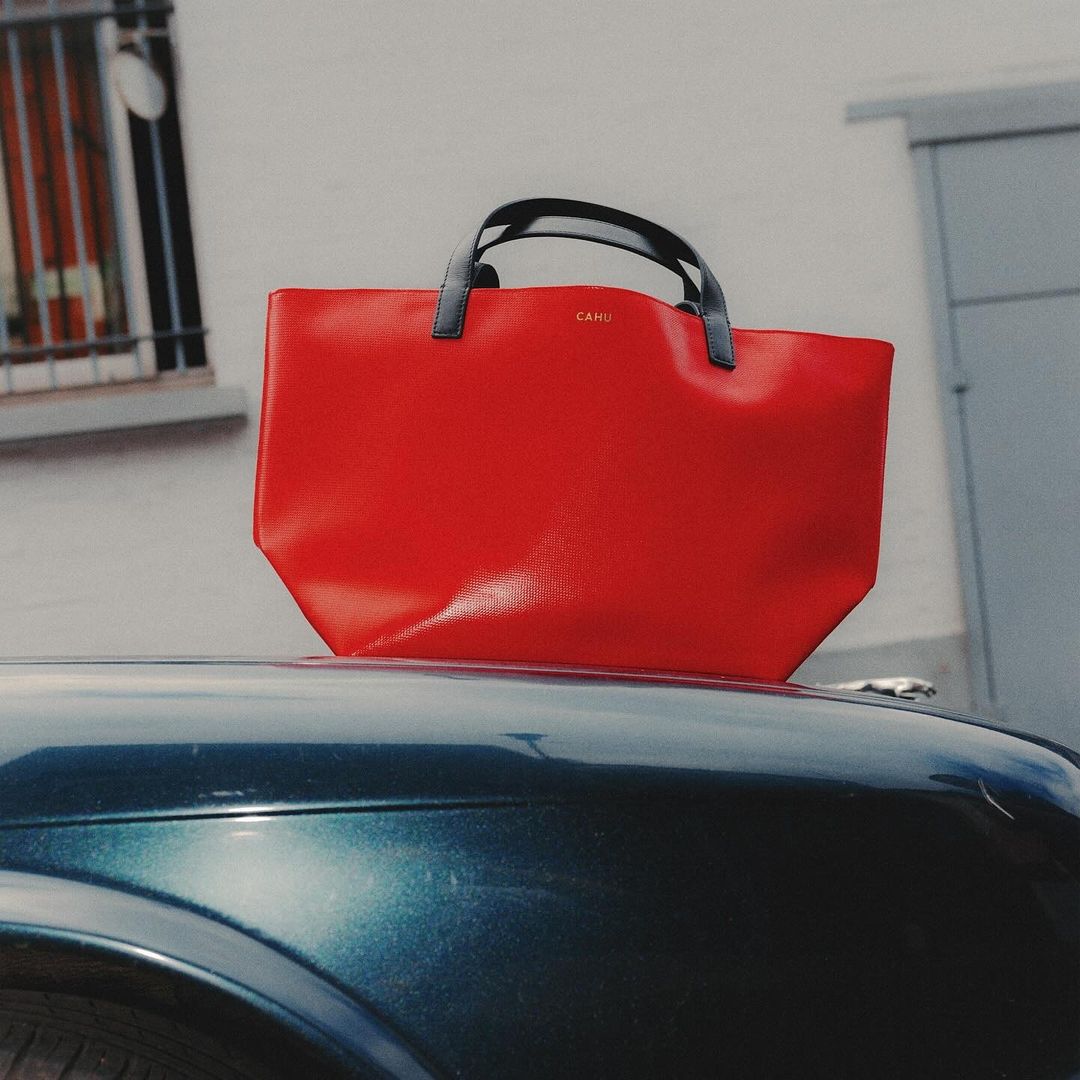 Red leather tote bag with black handles sitting on the hood of a blue-green car in front of a white building with a window.
