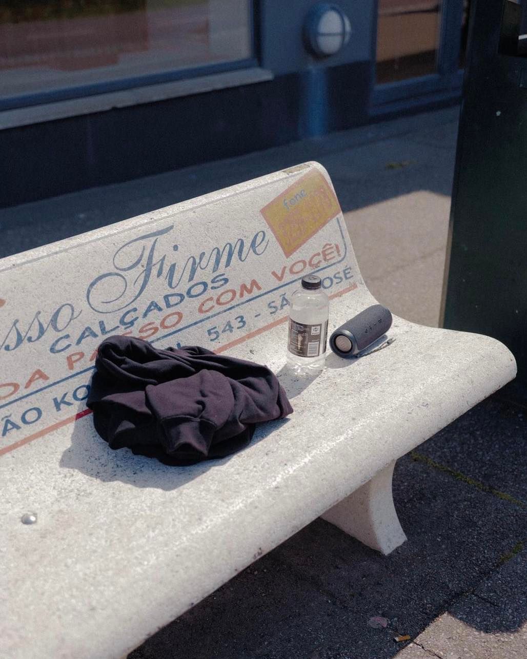 Outdoor shot of black clothing, a water bottle, and a speaker sitting on a stone bench with Portuguese writing, set against an urban backdrop.