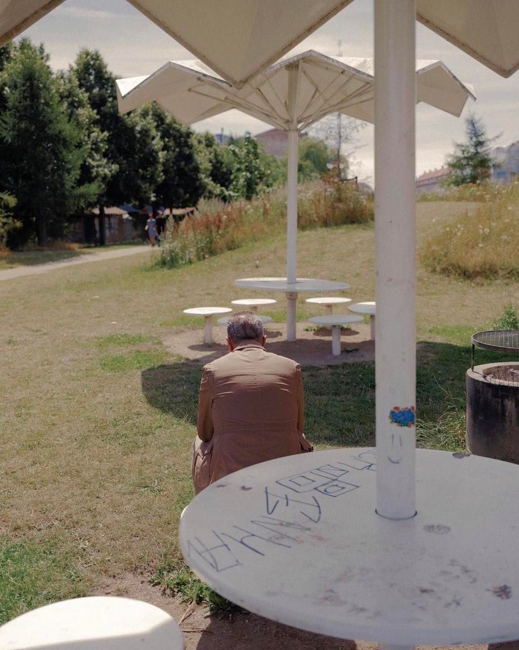 Man in jacket sits at a white table in a park under white umbrella structures with green trees, grass, and picnic tables in background.