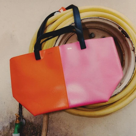 Two-tone pink and orange tote bag with black handles hangs on a wall with a yellow hose and faucet, shot in a studio with soft lighting.