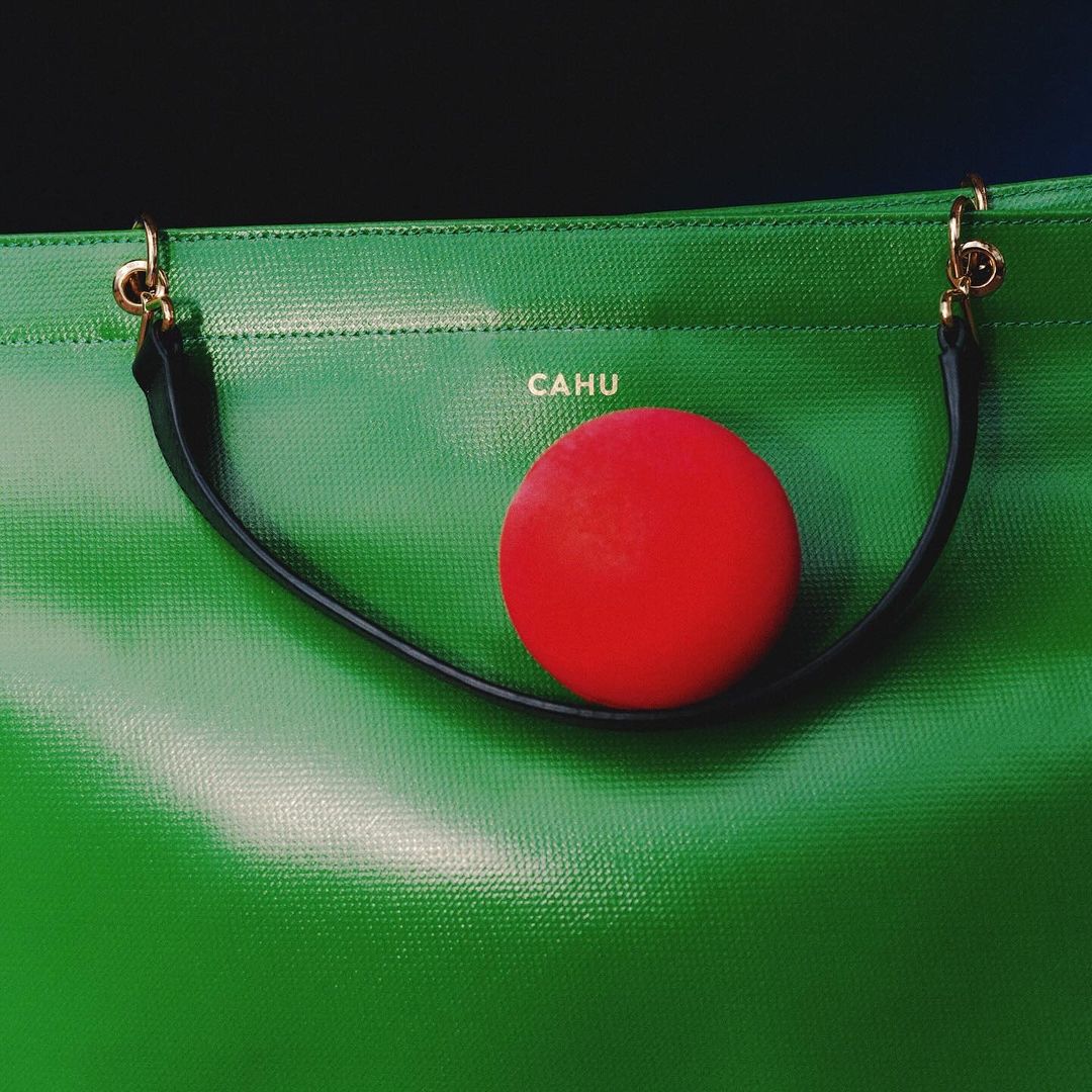A green CAHU bag with black handles is paired with a bright red round object against a dark background, creating a striking color contrast.