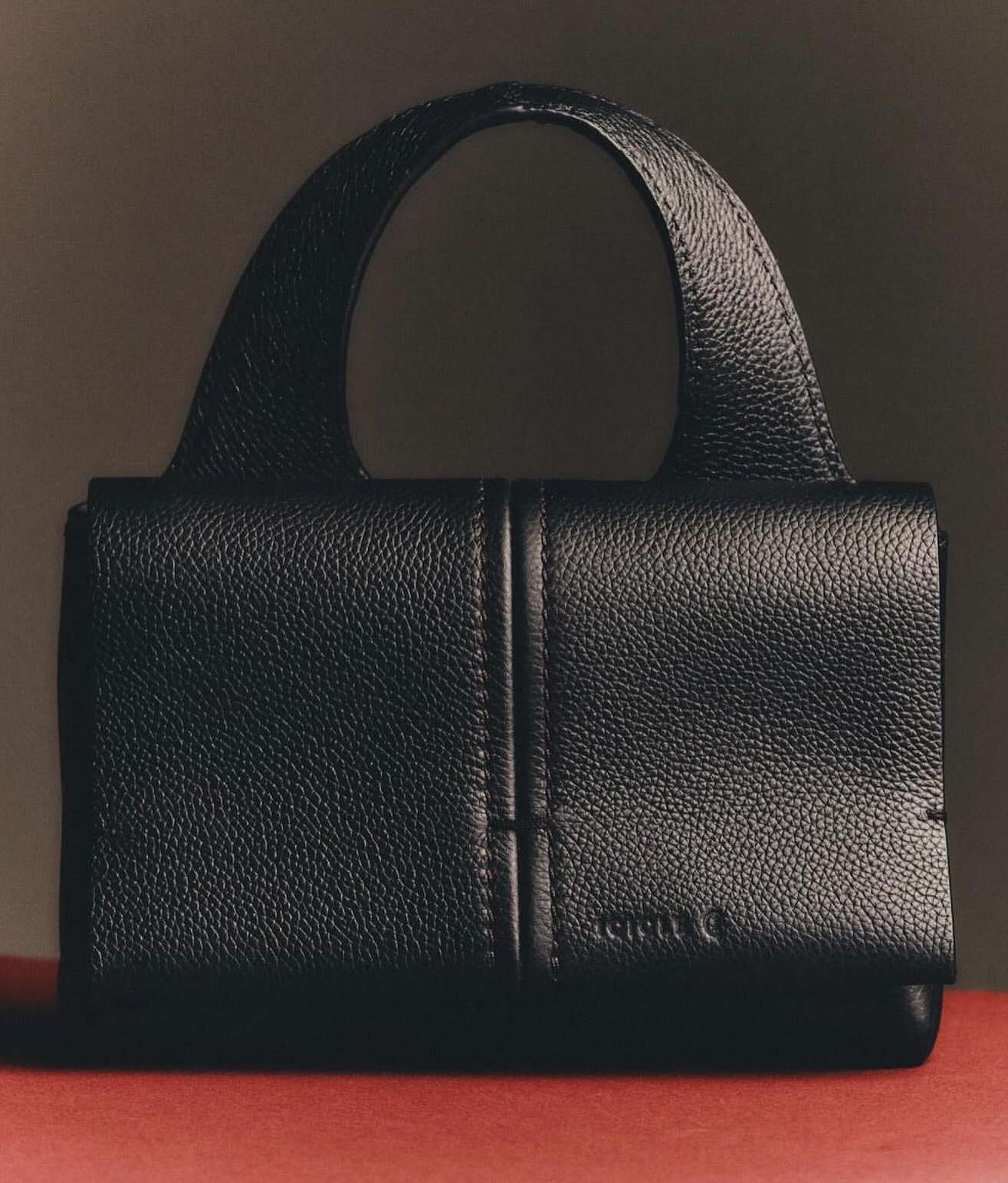 Close-up shot of a stylish black leather handbag featuring a top handle, sleek design, and subtle brand logo against a warm-toned background.