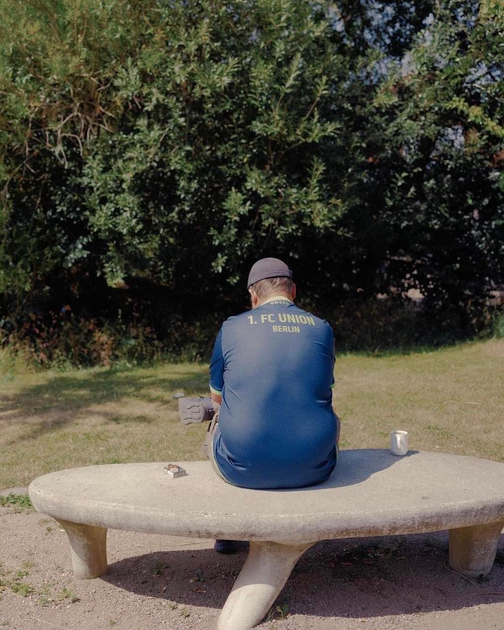 Man in soccer jersey sits on stone bench in park under trees on a sunny day.  He is wearing a hat, and the back of the jersey is visible.