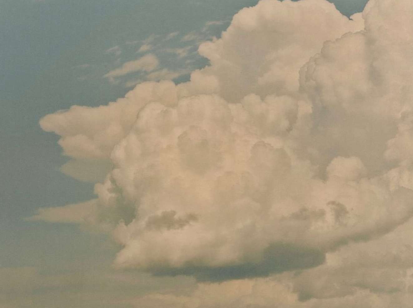 Dramatic cloudscape featuring dense, puffy clouds in the foreground, set against a pale blue sky. Soft light illuminates the cloud formations.