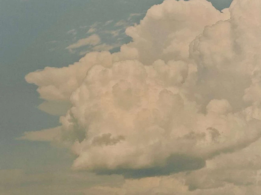 Dramatic cloudscape featuring dense, puffy clouds in the foreground, set against a pale blue sky. Soft light illuminates the cloud formations.