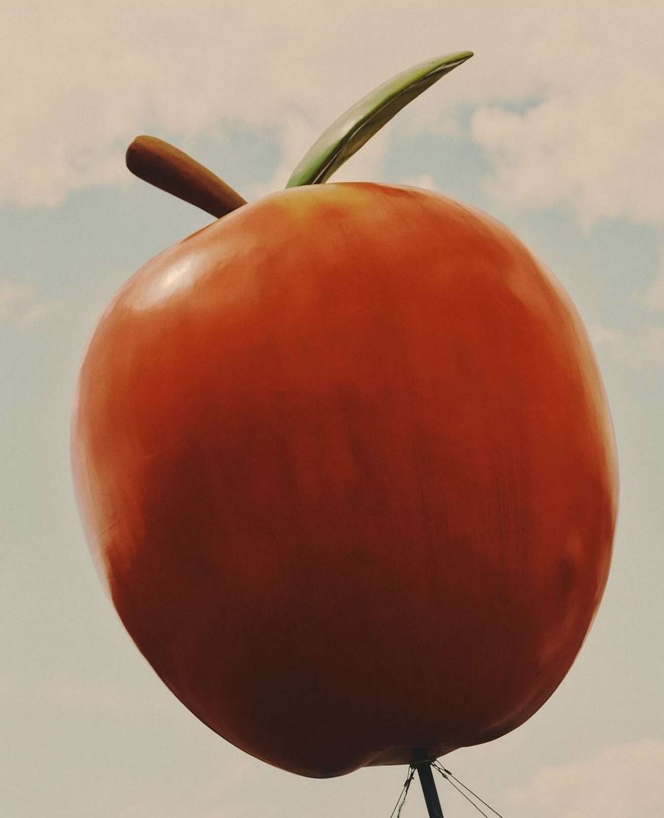An oversized roadside orange fruit with a green stem set against a light blue sky with clouds, designed as an attention-grabbing landmark.