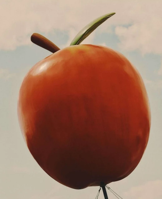 An oversized roadside orange fruit with a green stem set against a light blue sky with clouds, designed as an attention-grabbing landmark.