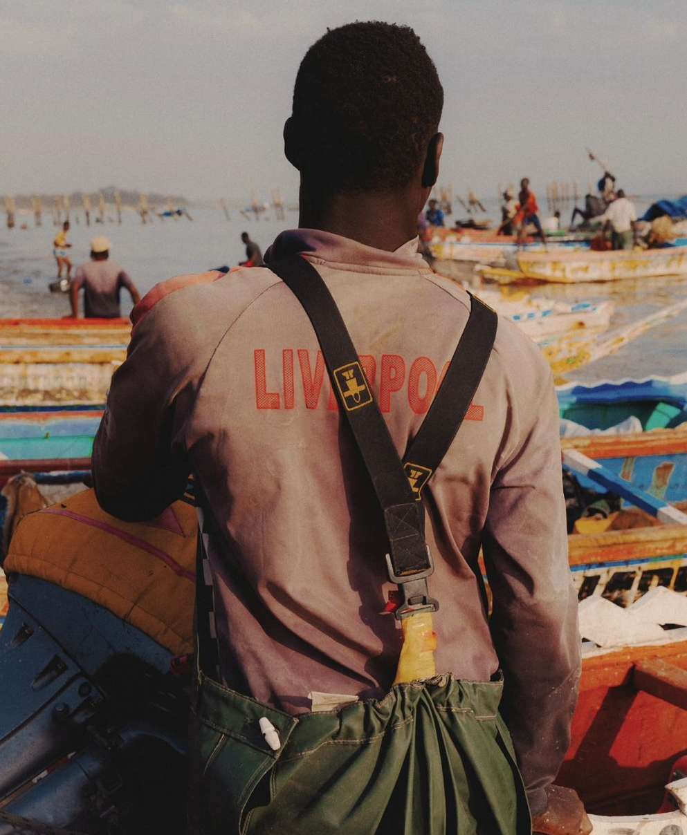 Rear view of dark-skinned adult male in work clothes, standing in a colorful fishing boat, overlooking a bay with people and boats.