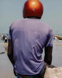 Man wearing a glossy orange helmet and purple t-shirt viewed from behind on a sandy beach.