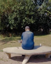 Man wearing a blue soccer jersey seen from behind seated on a stone bench outdoors with trees in the background