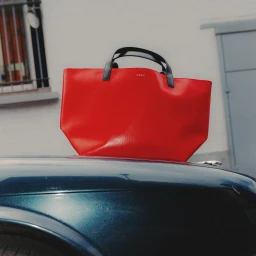 Bright red tote bag resting on the dark blue hood of a vintage car parked against a white wall.