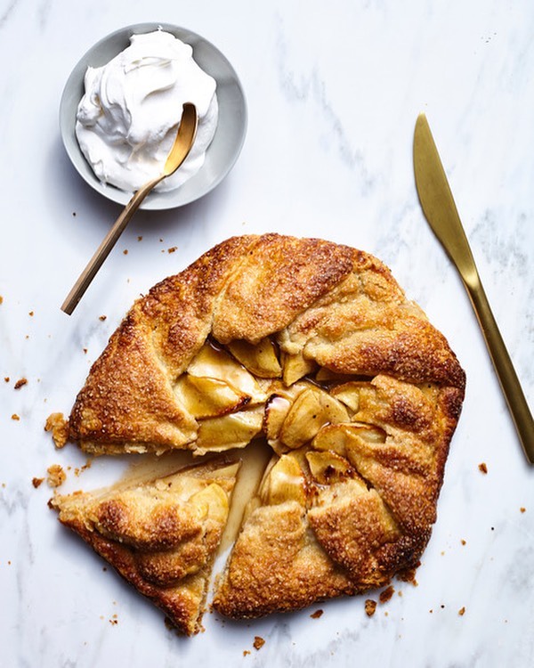 Overhead shot of an apple galette with golden crust, served with whipped cream and utensils on a white marble surface.