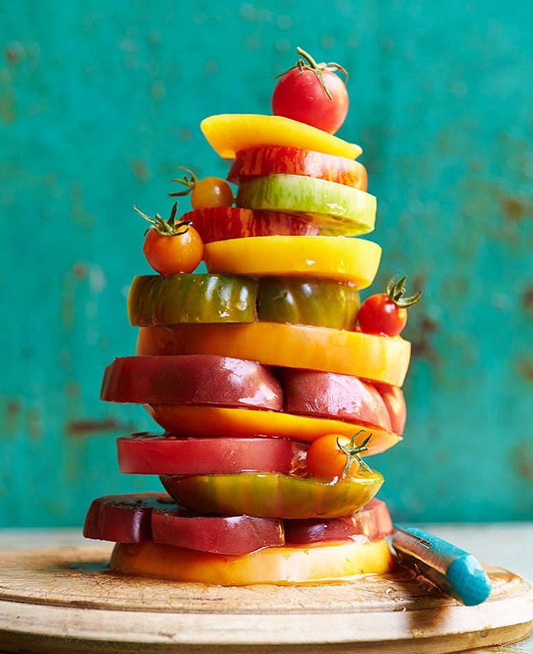 A stack of colorful sliced heirloom tomatoes and cherry tomatoes on a wooden board, set against a teal background.