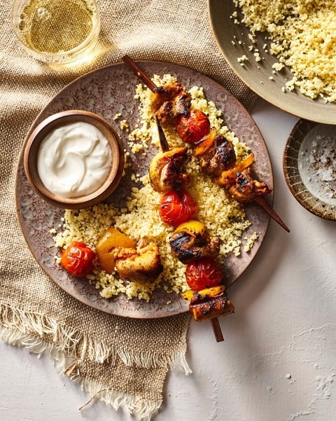 Overhead shot of chicken and vegetable skewers served with couscous, yogurt dip, and white wine on a rustic table setting.