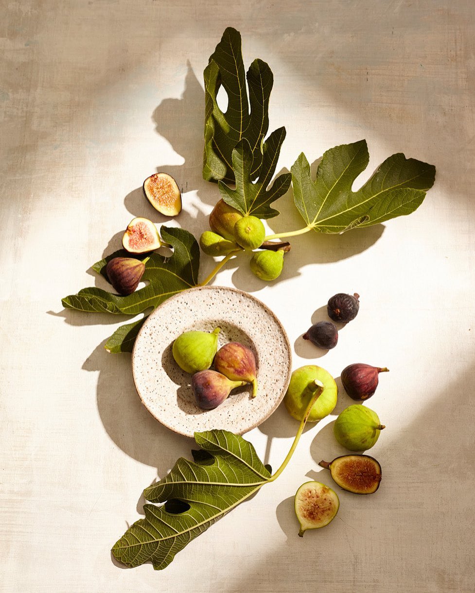 Fresh fig halves, whole figs, and fig leaves arranged on a white surface under natural lighting create an inviting, natural food still life.
