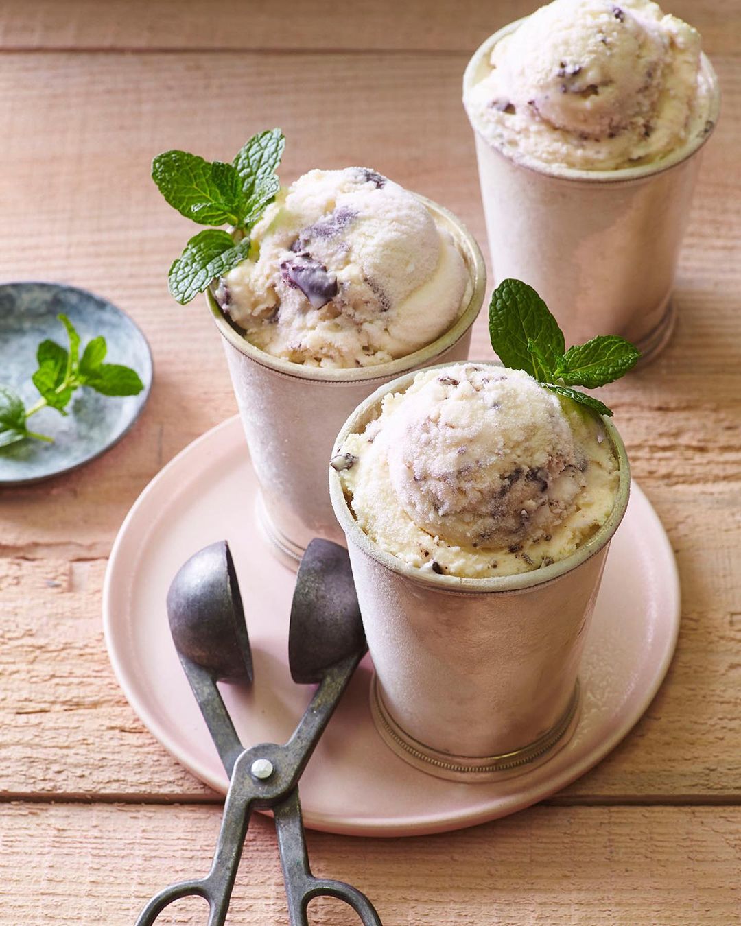Three servings of ice cream in metal cups with mint garnish, presented on a pink plate with antique ice cream scoop.
